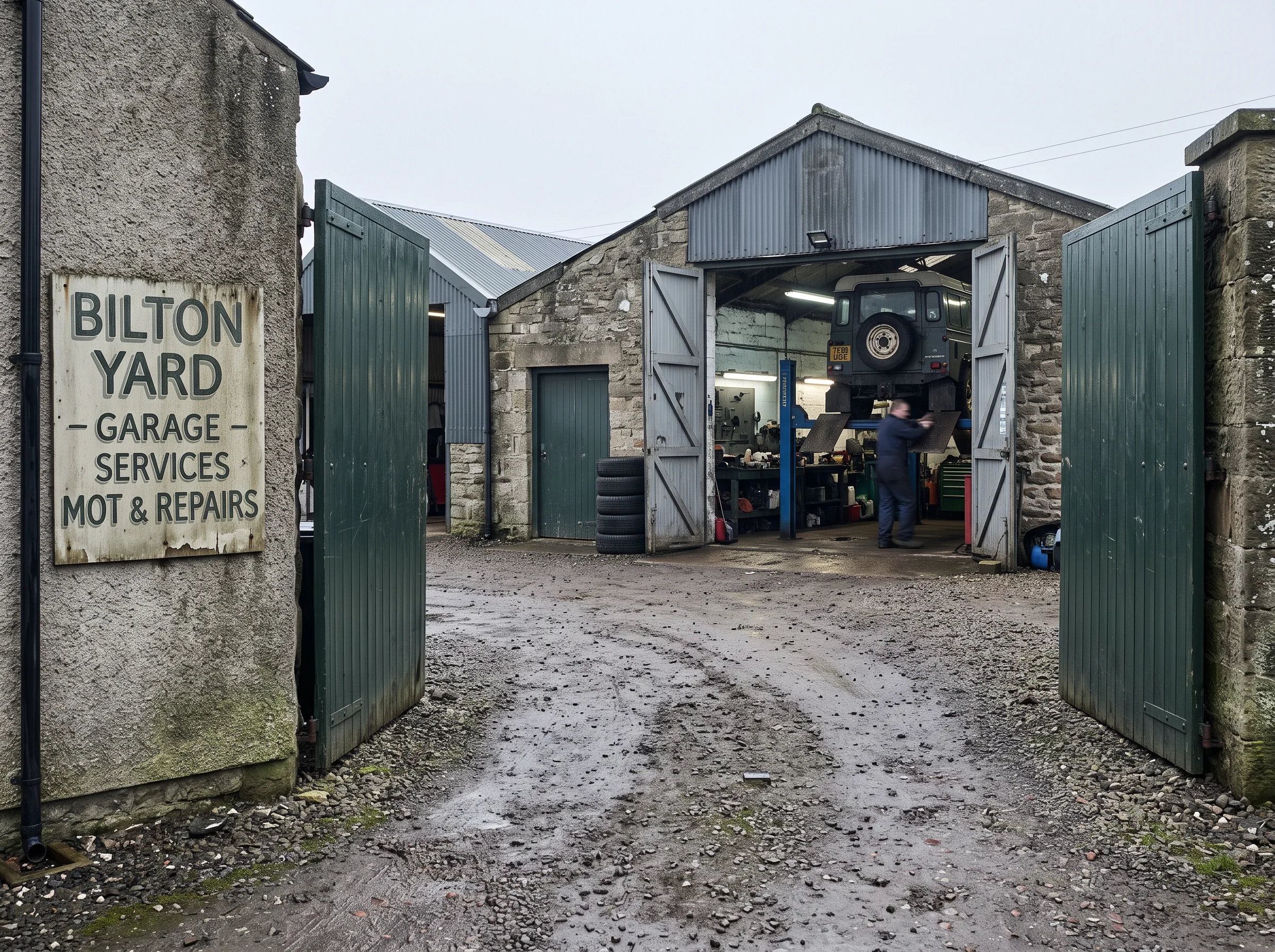 The yard gate from the road: gravel, a hand-painted name board, and open workshop doors beyond in flat grey light